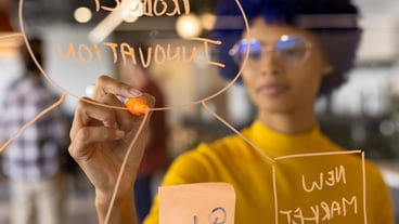 Biracial casual businesswoman with blue afro making notes on glass wall in office. Casual office, brainstorm, business and creative work, unaltered.