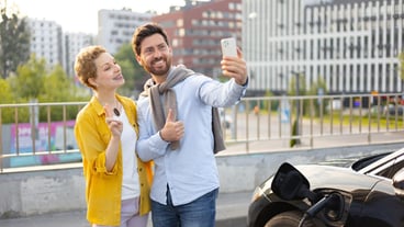 A smiling couple takes a selfie next to their electric vehicle while it charges highlighting the