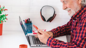 Portrait of an elderly man in a checkered shirt sitting at work with a laptop and smartphone in hand