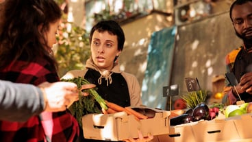 Young woman small business owner holding cardboard box full of fresh organic locally grown fruits and vegetables, female vendor in apron selling healthy pesticide free veggies at farmers market
