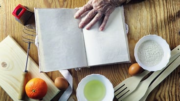 Open recipe book in the hands of an elderly woman in front of a table with utensils