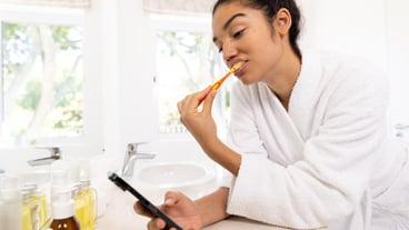 Biracial woman in bathrobe brushing teeth and using smartphone in sunny bathroom