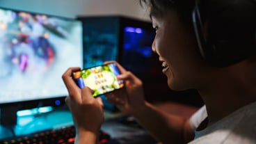 Photo of joyous gamer boy playing video games on mobile phone and computer in dark room, wearing headphones and using backlit colorful keyboard