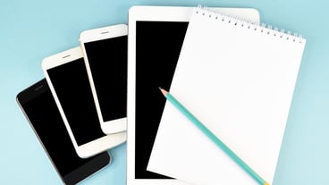 A notebook with a pencil lies on top of smartphones and a tablet computer
