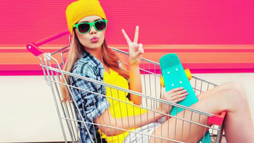Cool young smiling woman having fun sitting in trolley with skateboard on colorful pink background