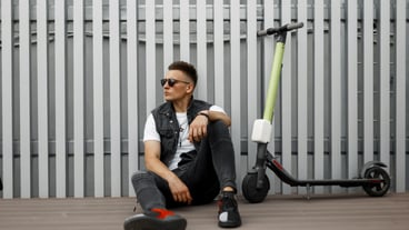 Stylish nice young hipster man in dark sunglasses in trendy summer clothes resting after riding an electric scooter