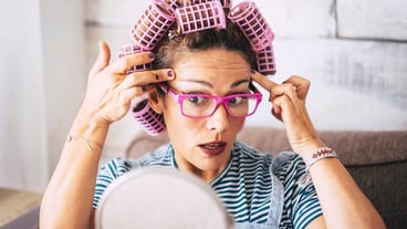 Young woman checking wrinkles near eye while applying curlers on hair and looking in front on mirror sitting on sofa in living room at home. Beautiful woman in eyeglasses getting ready with make up.
