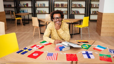 Cheerful young man sitting at a table with books and country flags in the library