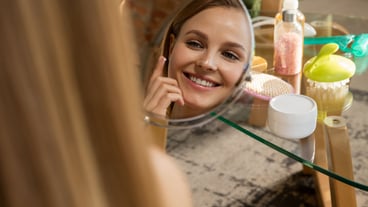 Beauty Day. Woman wearing towel doing her daily skincare routine at home. Putting on moisturizing and creme, looking on her reflection in mirror. Concept of beauty, self-care, cosmetics, youth.