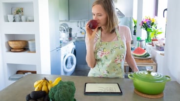Thoughtful woman smelling fruit while cooking in her kitchen, with tablet near pot and fresh vegetables on the counter. Front view. Home cooking and healthy eating concept