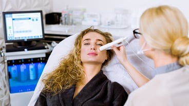 An esthetician performs ultrasonic facial cleansing and pulse therapy on a pretty female client seated in a chair wearing a grey coat at a beauty spa clinic.