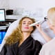 An esthetician performs ultrasonic facial cleansing and pulse therapy on a pretty female client seated in a chair wearing a grey coat at a beauty spa clinic.
