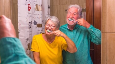 Couple of two happy and smiling seniors brushing their teeth of they each other together at home in the bathroom - self care and taking care of theyself