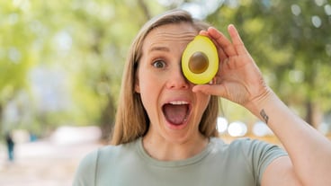 Young blonde woman at outdoors holding an avocado with surprised expression