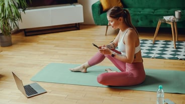 Young woman rests her thigh and use mobile phone sitting on a yoga mat in front of a laptop