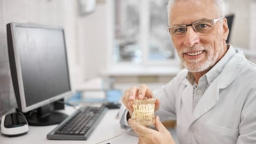 Cheerful elderly dentist holding a full jaw dental implant while smiling at the camera