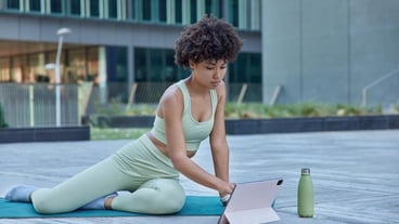 Slim active woman practices yoga uses digital tablet for having online training watches tutorial drinks fresh water dressed in activewear poses outdoors against blurred background. Fitness concept