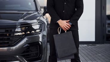 Close up view of businessman in black suit and tie stands near modern automobile with shopping bags in hands.