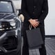 Close up view of businessman in black suit and tie stands near modern automobile with shopping bags in hands.