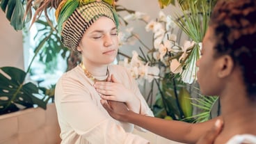 Bioenergy. Dark-skinned and caucasian young adult woman with closed eyes sitting opposite each other touching hand meditating in spa salon