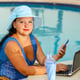 A young woman in a hat at a table in the morning by the pool with a smartphone and works remotely