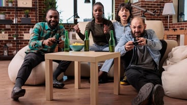 Happy friends laughing while they play strategy video games, using controller and tv console to have fun together. Young people enjoying leisure activity with beer bottles and gaming teamwork.