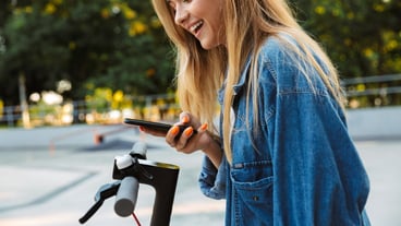 Happy positive smiling cheerful teenage girl in park walking on scooter using mobile phone.