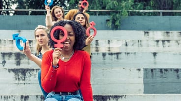 A Black woman jokes with a female cork symbol in the foreground as more people do the same behind her sitting on a flight of stairs.