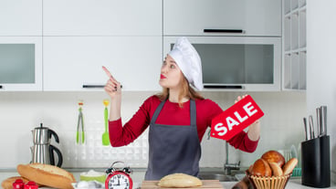 Front view blonde woman in chef's hat and apron with red sale sign in the kitchen.