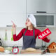 Front view blonde woman in chef's hat and apron with red sale sign in the kitchen.