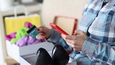 Woman sitting on the sofa with a box of shoes, holding a bank card and a smartphone.