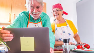 Couple of two happy seniors cooking together in the kitchen learning and following a video tutorial of cook in the laptop -  preparing healthy food together