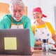 Couple of two happy seniors cooking together in the kitchen learning and following a video tutorial of cook in the laptop -  preparing healthy food together