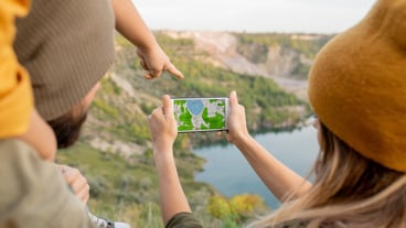 A little boy's hand points to a smartphone screen with a map held by his mother, who is standing by a river and deciding where to go next.