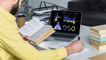 bearded man studying with book near laptop with charts and graphs on screen in modern office