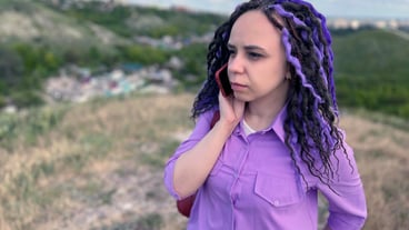 Young beautiful woman talking on mobile phone standing in hilly terrain Cute vivid female with dreadlocks speaking on smartphone standing outdoors in cloudy windy summer weather