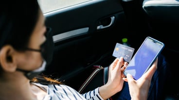 Young Asian woman sitting in the back seat of a car holding a credit card in her hand using a cell phone.