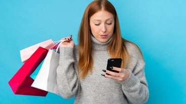 Young red-haired woman above isolated blue wall holding shopping bags and writing a message to a friend on her cell phone