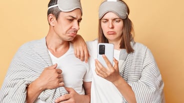 Sad unhappy couple man and woman in sleep eye mask wrapped in blanket isolated over beige background using mobile phone after waking up reading unpleasant news