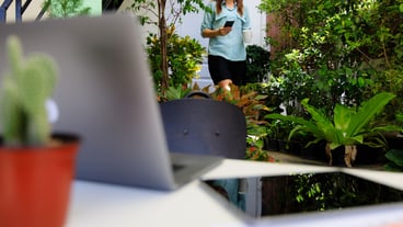 Businesswoman holding coffee cup and smartphone walking from home room to working desk in corner small garden with laptop and tablet computer. Concept of new normal people and work at home.
