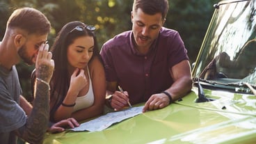 Young friends reading map that is on the hood of modern green jeep in the forest.