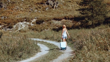 An attractive girl in shorts walks along a narrow summer path holding a travel suitcase in her hand