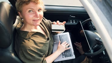 A middle-aged woman sits behind the wheel of a car with a laptop on her lap.