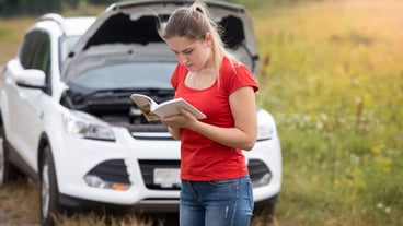 Portrait of young stressed woman standing at broken car and reading owner manual