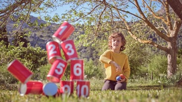 Positive boy kneeling on grassy meadow and throwing bags in pyramid of red tin cans while playing game in nature
