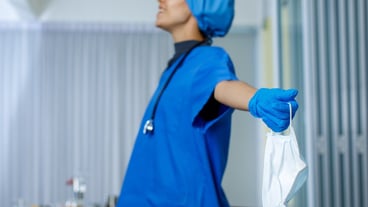 Closeup shot of used surgical face mask in hand of female happy smiling freedom doctor in blue hospital uniform rubber gloves and stethoscope in blurred background after coronavirus pandemic end.