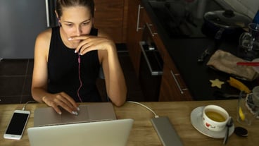 Young woman working on a laptop with a smart phone in her hands at home in the kitchen working remotely