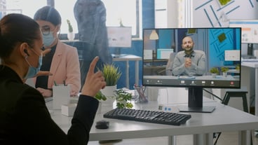 Freelancer wearing protective face mask during online video call sits at a desk in an office of a start-up company. Colleagues working on computer during a global coronavirus pandemic
