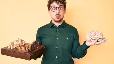 Young Caucasian with curly hair holding brain while playing chess puffing cheeks with funny face mouth inflated with air catching air.