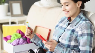 Smiling woman holding bank card and smartphone while sitting on couch with box of shoes online